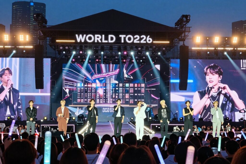 A vibrant concert scene showcasing BTS's world tour in 2026, with the stage framed in the foreground featuring elaborate lighting and vibrant visuals. The members are depicted in stylish yet modest performance attire, engaging with the enthusiastic crowd full of fans holding lightsticks, all illuminated by dazzling spotlights. In the middle ground, a large LED screen displays tour graphics, enhancing the excitement with a dynamic atmosphere. The background features a city skyline silhouetted against a twilight sky, adding depth and context to the scene. The overall mood is electric and full of energy, capturing the spirit of a legendary concert night. The focus is sharp, with a slight bokeh effect on the audience to emphasize the performers.