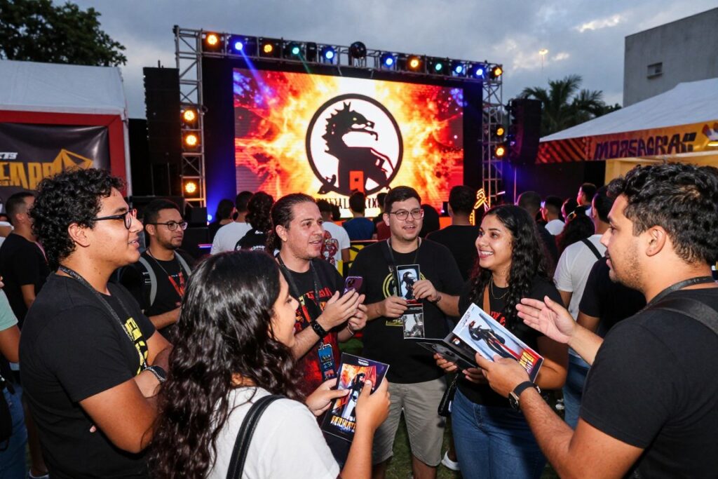 A vibrant and dynamic scene depicting a cinematic release event for "Mortal Kombat 2" in Brazil. In the foreground, excited fans dressed in casual gaming attire gather to discuss the game, showing a mix of anticipation and joy. Some hold collectible posters and gaming memorabilia. In the middle ground, a large screen displays the iconic Mortal Kombat 2 logo, with colorful lights illuminating the surroundings, creating an energetic atmosphere. The background features a bustling gathering space adorned with themed decorations and banners, capturing the essence of the gaming culture. The scene is set during the evening with soft yet vibrant lighting to enhance the mood, shot from a slightly elevated angle to encompass the vibrant crowd. The image should evoke excitement and community spirit, reflecting a celebratory launch atmosphere.