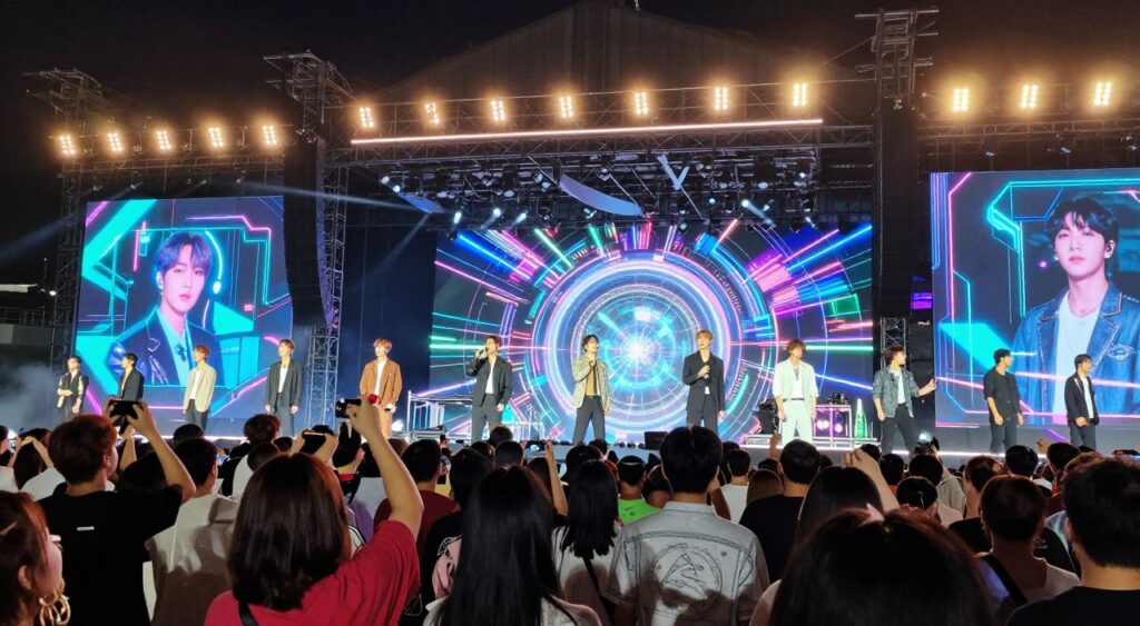 A dynamic concert scene featuring a technologically advanced BTS show, showcasing stunning stage designs with holograms and vibrant LED displays. In the foreground, a diverse crowd of fans, dressed in casual, colorful attire, are captivated by the live performance. The middle ground includes the BTS members in stylish, professional performance outfits, engaging with the technology around them. The background reveals a futuristic stage setup with intricate light patterns and machinery, illuminating the night sky. The atmosphere is electric, filled with excitement and wonder. Use dramatic lighting to emphasize the powerful visuals, shot from a low angle to capture the scale of the performance and the awe on the audience's faces.